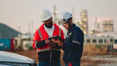 two men are standing next to a car looking at a tablet .